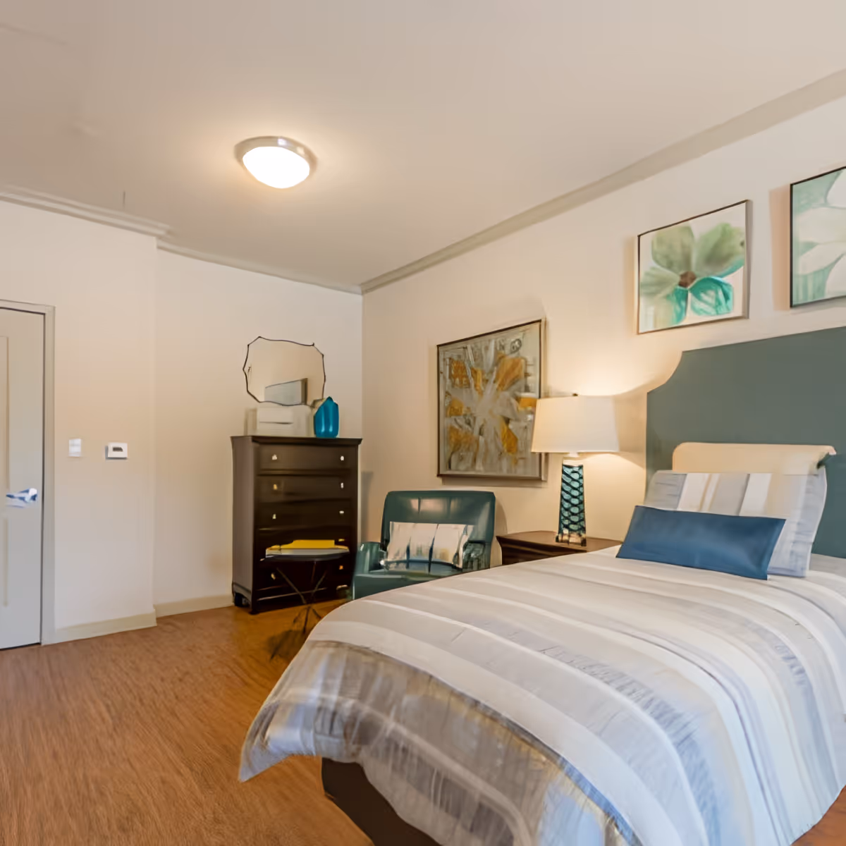 A neatly made bed with striped bedding and a blue accent pillow in a bedroom. Next to the bed is a nightstand with a lamp. A teal armchair with a decorative pillow is placed near the nightstand. On the wall above the bed are two framed floral artworks, and another framed abstract painting hangs on the adjacent wall. A dark wooden dresser with a mirror and decorative items on top is positioned against the far wall. The room has light-colored walls and a ceiling light fixture.