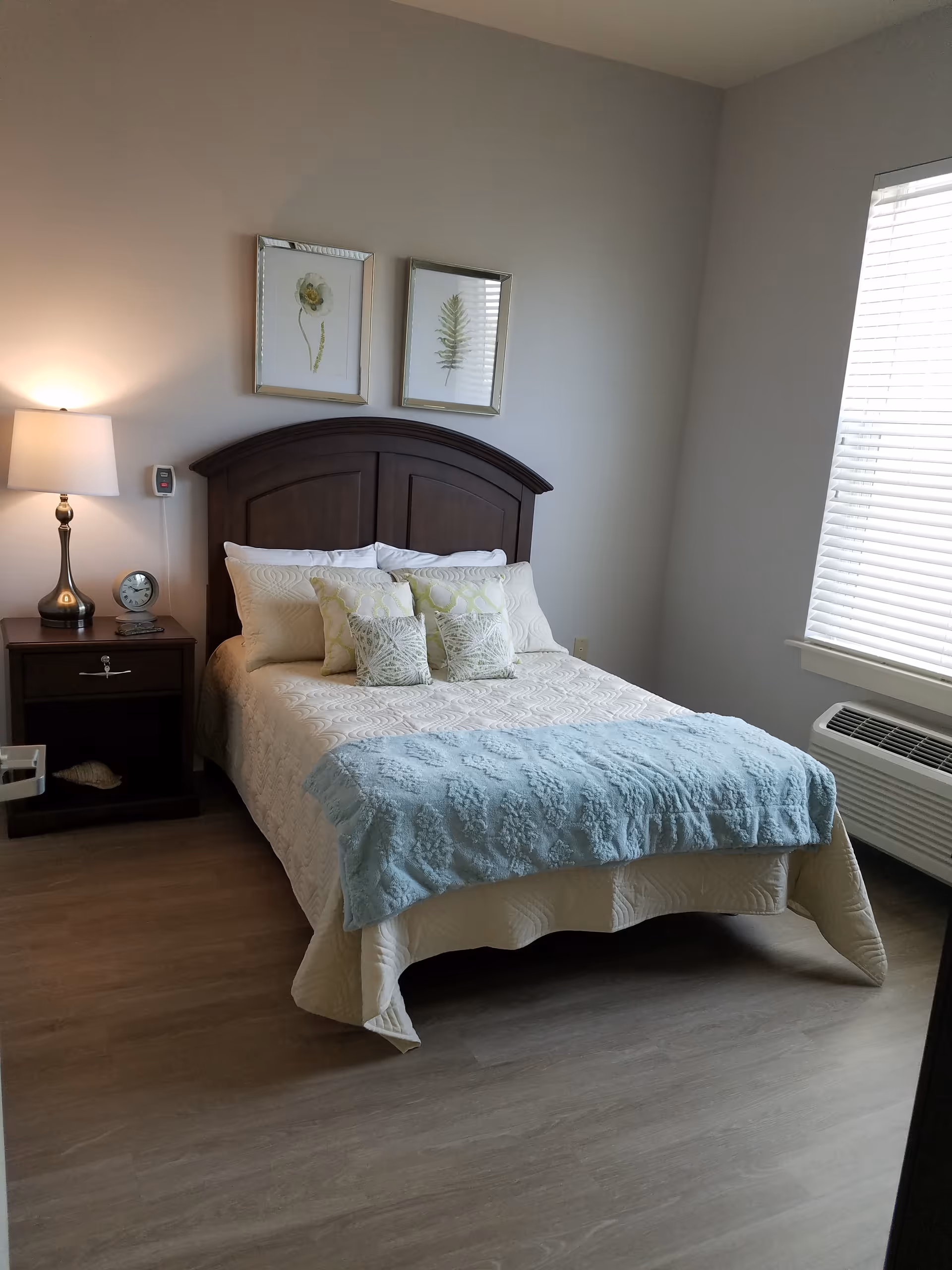 A neatly made bed with a wooden headboard in a bedroom. The bed has white bedding with a light blue throw blanket at the foot and several decorative pillows. There is a nightstand with a lamp and a clock on the left side of the bed. Two framed botanical prints hang on the wall above the headboard. A window with blinds is on the right side of the room, and an air conditioning unit is below the window.