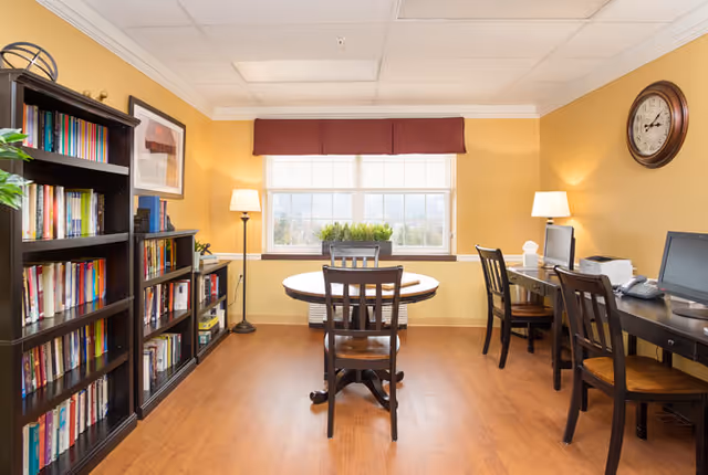 A cozy room with yellow walls and wooden flooring featuring a round table with four chairs in the center. On the left side, there are two black bookshelves filled with books and decorative items. On the right side, there are two desks with chairs, desktop computers, and lamps. A large window with a red valance lets in natural light, and a round wall clock is mounted on the right wall.