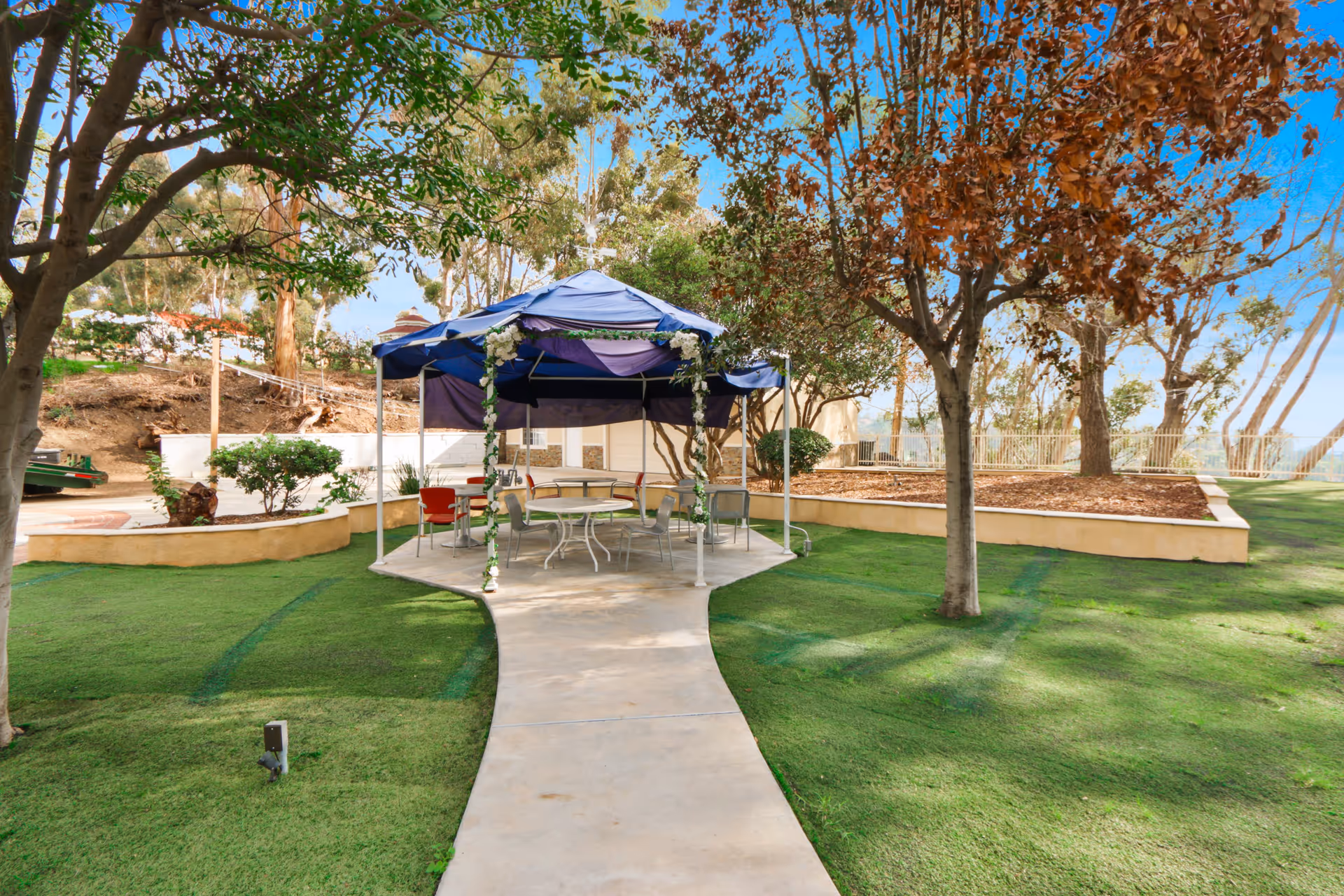 Outdoor garden area with a paved walkway leading to a blue canopy tent decorated with white flowers. Under the tent are several chairs and round tables. The surrounding area has green grass, trees with some brown leaves, and a retaining wall with mulch and bushes.