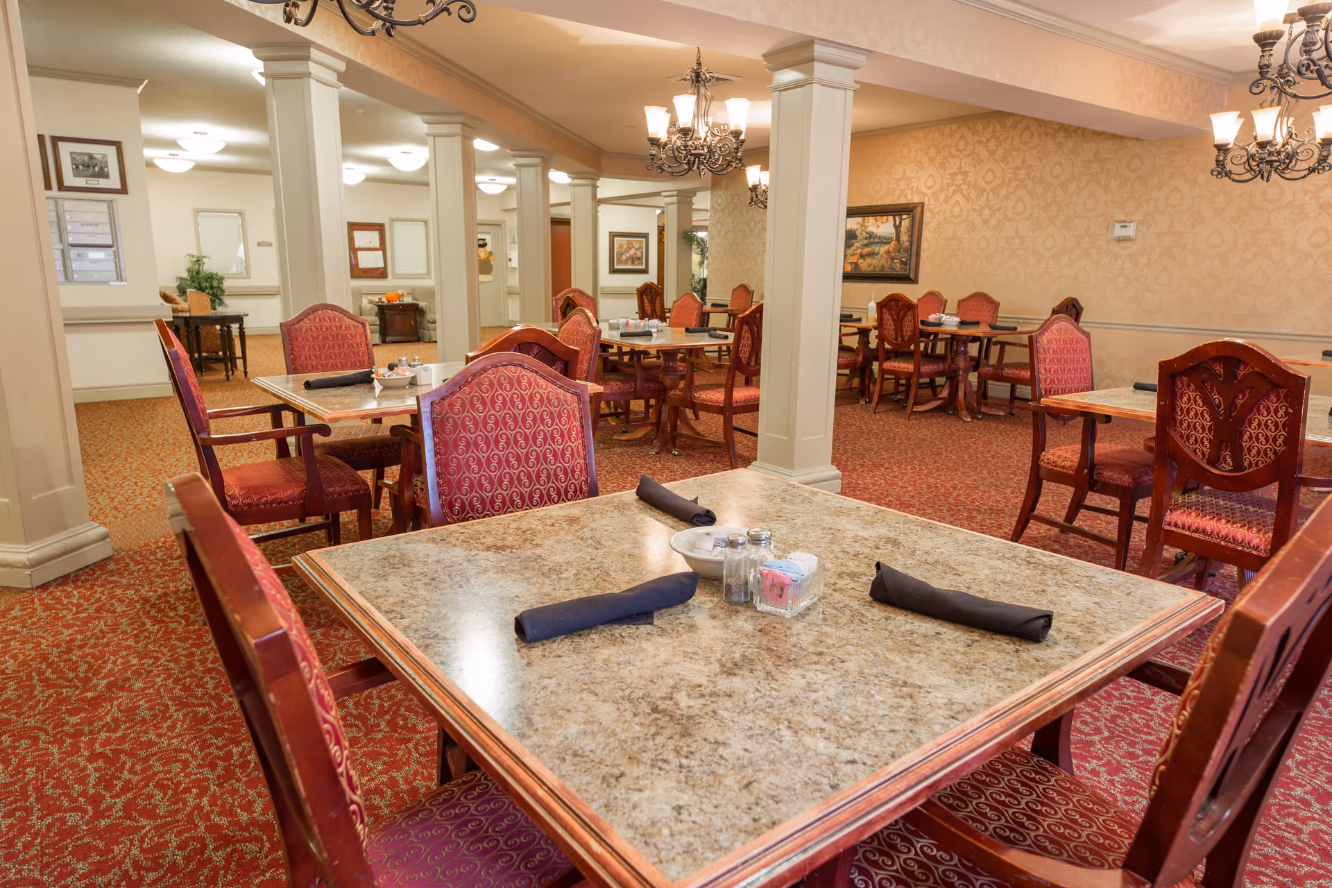 A dining room in a senior living facility with multiple tables and red upholstered chairs. The tables have marble-like surfaces and are set with black napkins, salt and pepper shakers, and small condiment containers. The room features beige patterned wallpaper, red carpet with a floral design, white columns, and ornate chandeliers hanging from the ceiling. There are framed pictures on the walls and a hallway visible in the background.