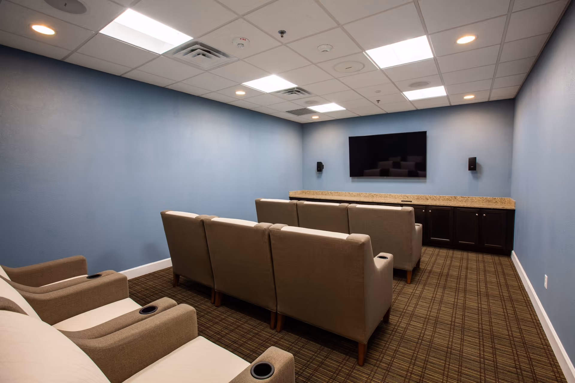 Small media/theater room with rows of upholstered recliner seats facing a wall-mounted flat-screen TV.
