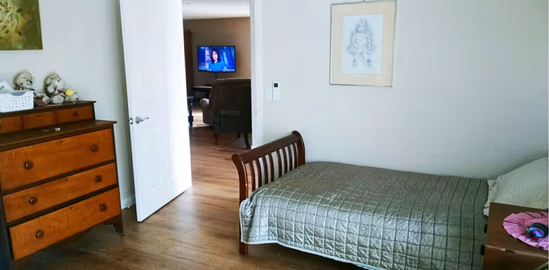 A bedroom with a single wooden bed covered with a gray quilt. To the left is a wooden dresser with decorative items on top. The door is open, showing a glimpse of a living room with a television and armchairs. A framed sketch hangs on the wall above the bed.