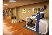 Interior view of a rehabilitation or physical therapy area in a senior living facility with wooden flooring, a treadmill machine, exercise equipment, and framed pictures on the wall.