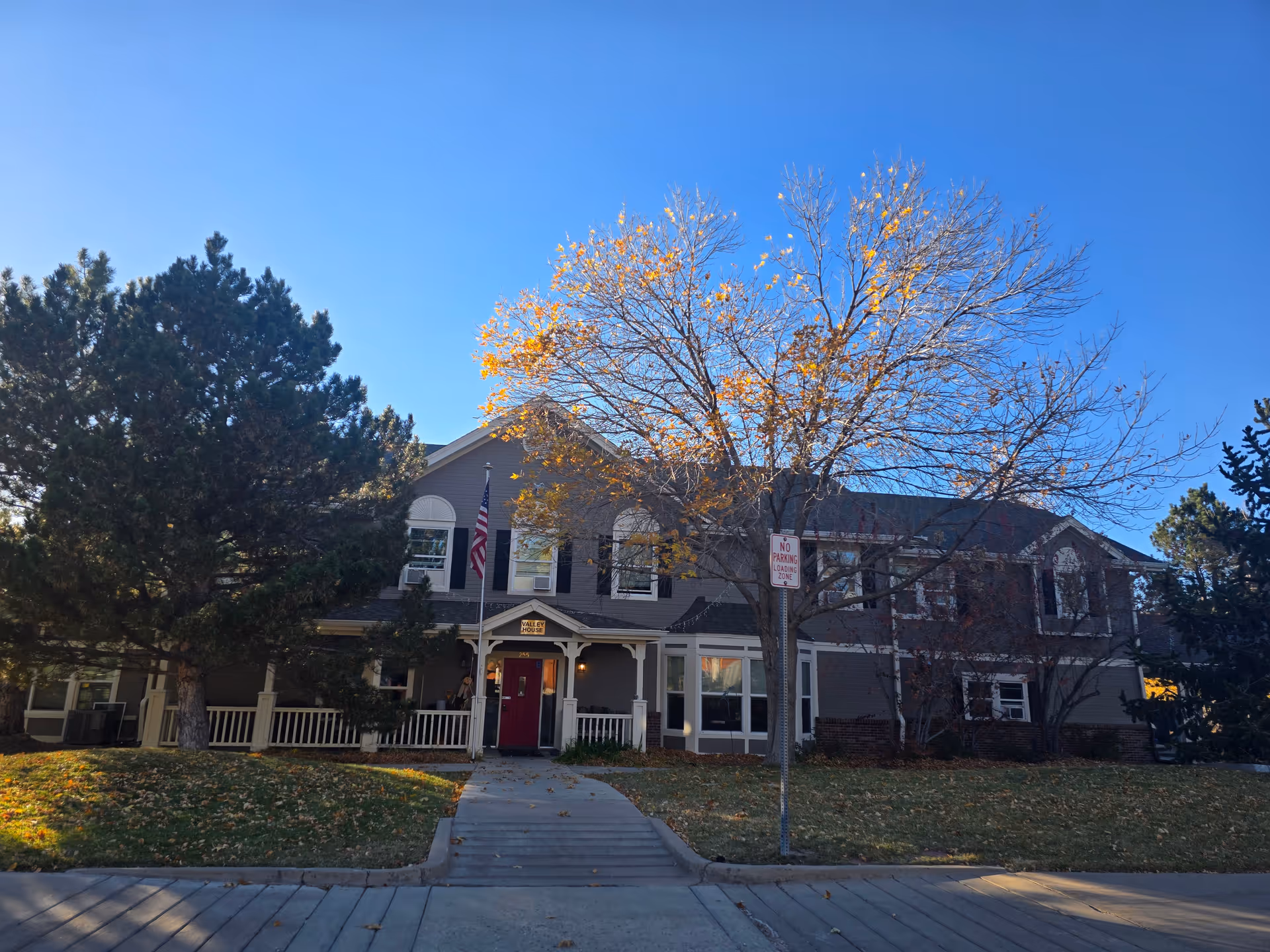 Exterior view of a two-story assisted living facility building with a front porch, an American flag, and a tree with autumn leaves in front. The sky is clear and blue.