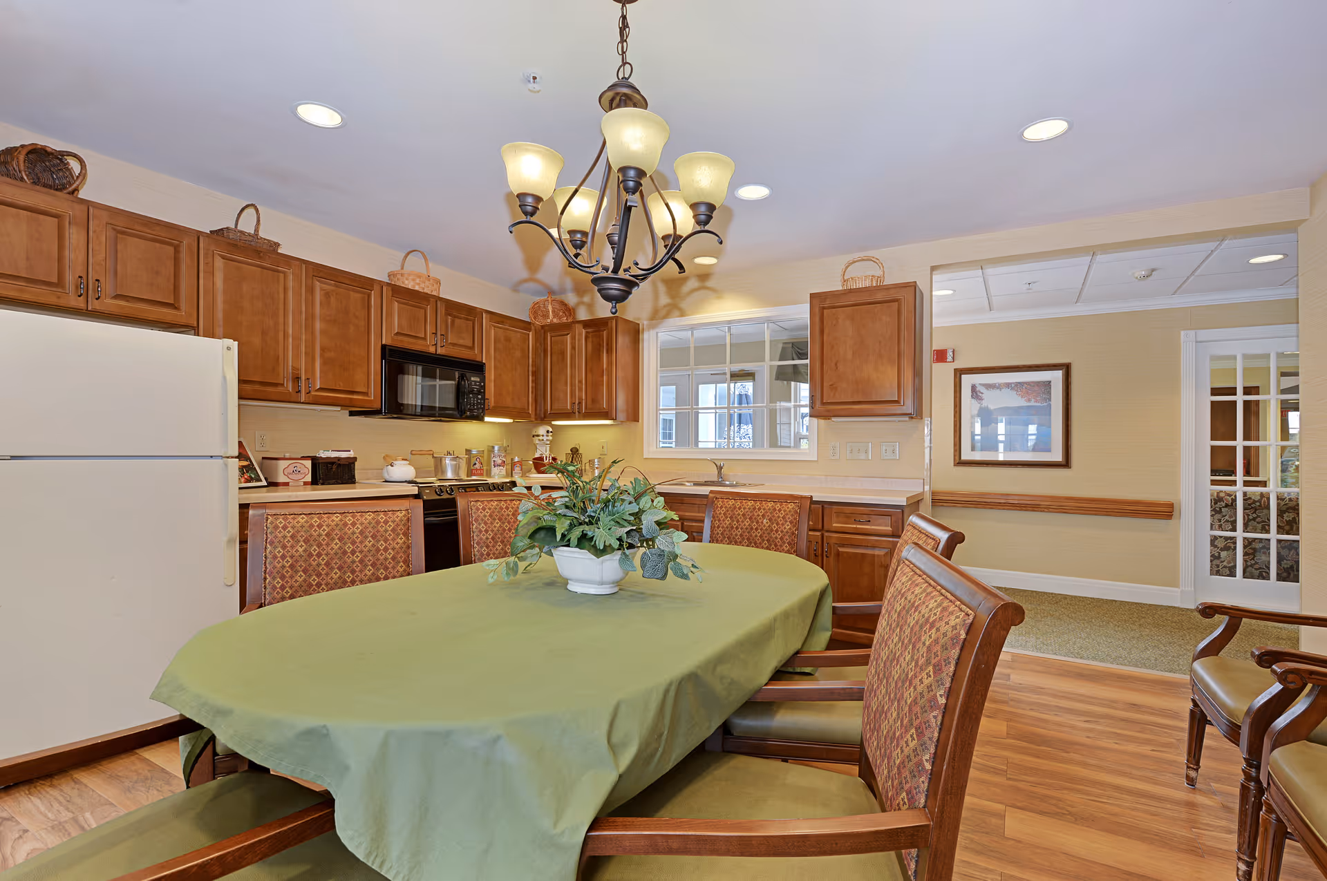 Dining table with a green tablecloth and centerpiece in a communal dining area adjoining a kitchen with wood cabinets and appliances.