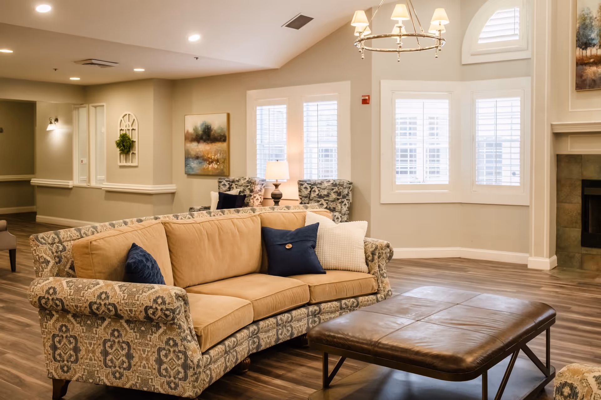 A cozy living room area in a senior living facility featuring a patterned sofa with beige cushions and blue pillows, a large leather ottoman, two patterned armchairs near windows with white shutters, a table lamp, a chandelier, and a fireplace with a painting above it. The room has wooden flooring and neutral-colored walls.