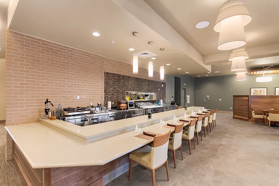 A modern dining area with a long counter featuring multiple chairs set with placemats and napkins. Behind the counter is a kitchen area with stainless steel appliances and cooking equipment. The room has pendant lights hanging from the ceiling and a tiled floor, with additional seating booths visible in the background.