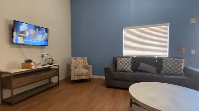 A living room with a mounted flat-screen TV on the wall, a wooden console table beneath it with some electronic devices and cables, a patterned armchair, a dark gray sofa with three patterned cushions, and a round white coffee table. The walls are painted blue and white, and there is a window with closed blinds.