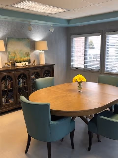 Round wooden dining table with teal upholstered chairs and a vase of yellow flowers in a bright room with a sideboard, lamps, and windows.
