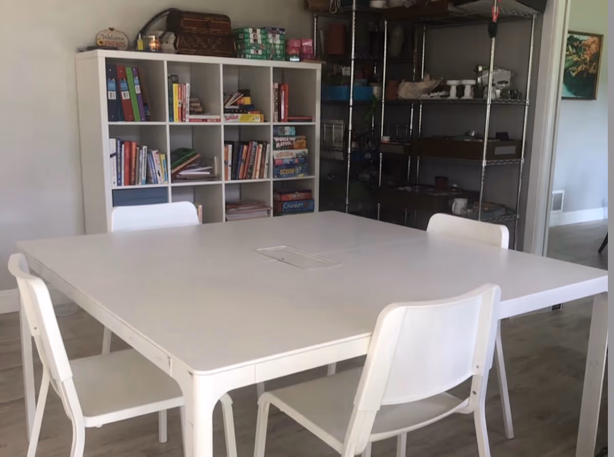 A room with a white square table surrounded by four white chairs. Behind the table is a white cubby bookshelf filled with books and board games. To the right, there is a metal shelving unit with various items and a doorway leading to another room.
