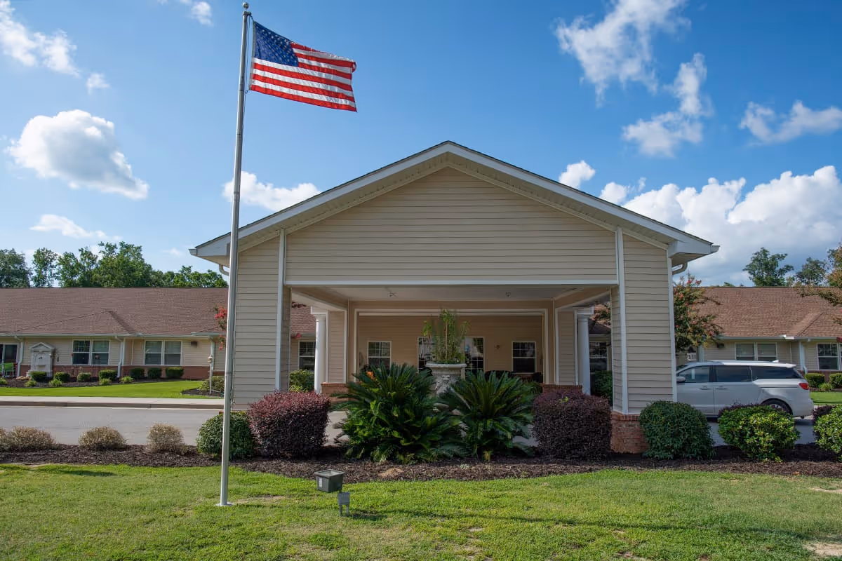 Exterior view of the entrance to Spring Oak at Lexington facility with a covered driveway, landscaped bushes, an American flag on a flagpole, and a blue sky with scattered clouds.
