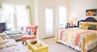 Bright and cozy bedroom with a large bed covered in a colorful patchwork quilt, a yellow coffee table with a bowl of fruit, a plaid armchair, a small TV on a stand, and a door leading outside. The room is decorated with light curtains and a floral painting on the wall.