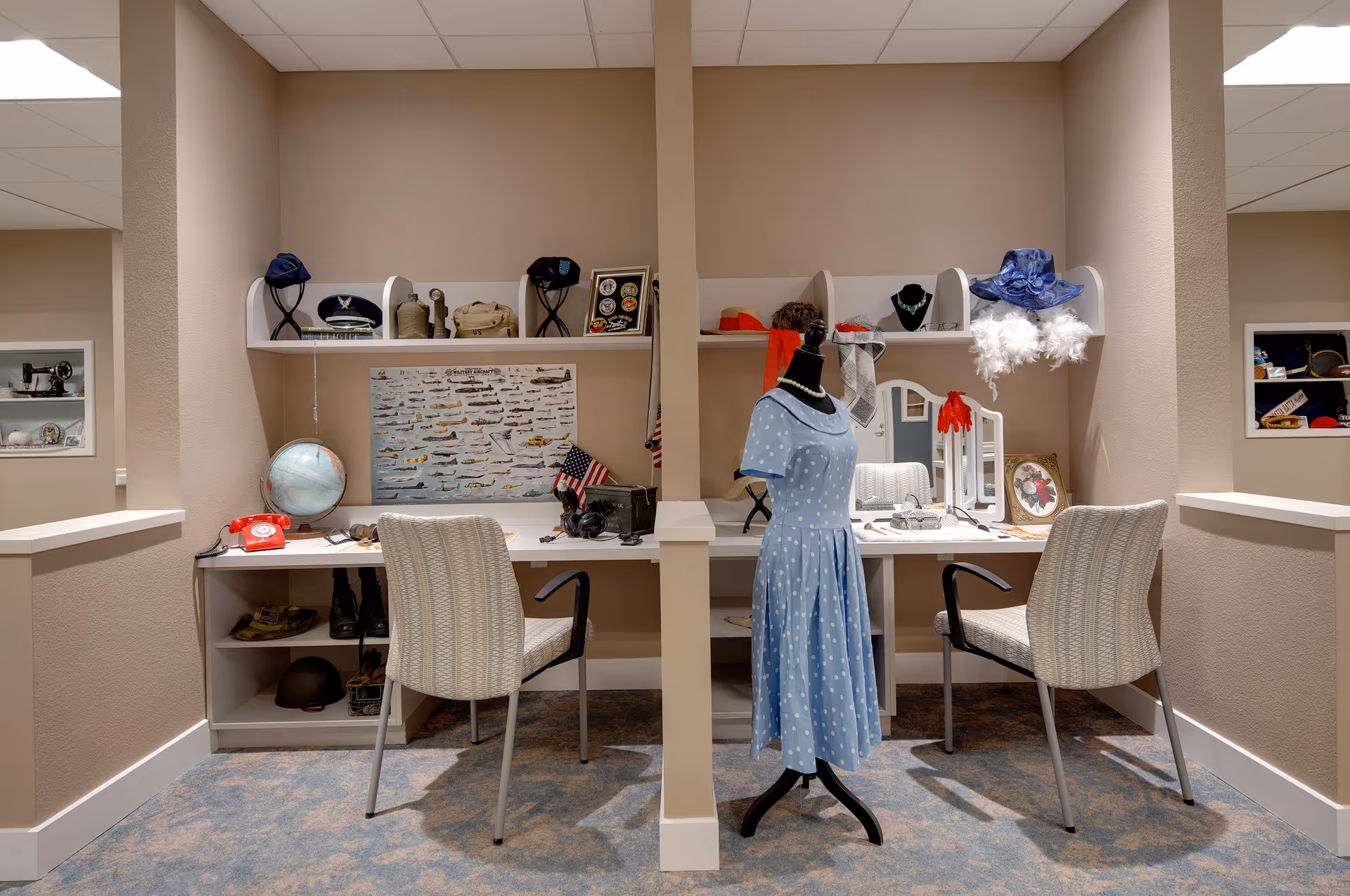 A cozy interior room with two workstations separated by a partial wall. Each workstation has a chair and shelves above holding various vintage items including hats, a globe, and framed pictures. A mannequin dressed in a blue polka dot dress stands in the center. The room has beige walls and a patterned carpet.