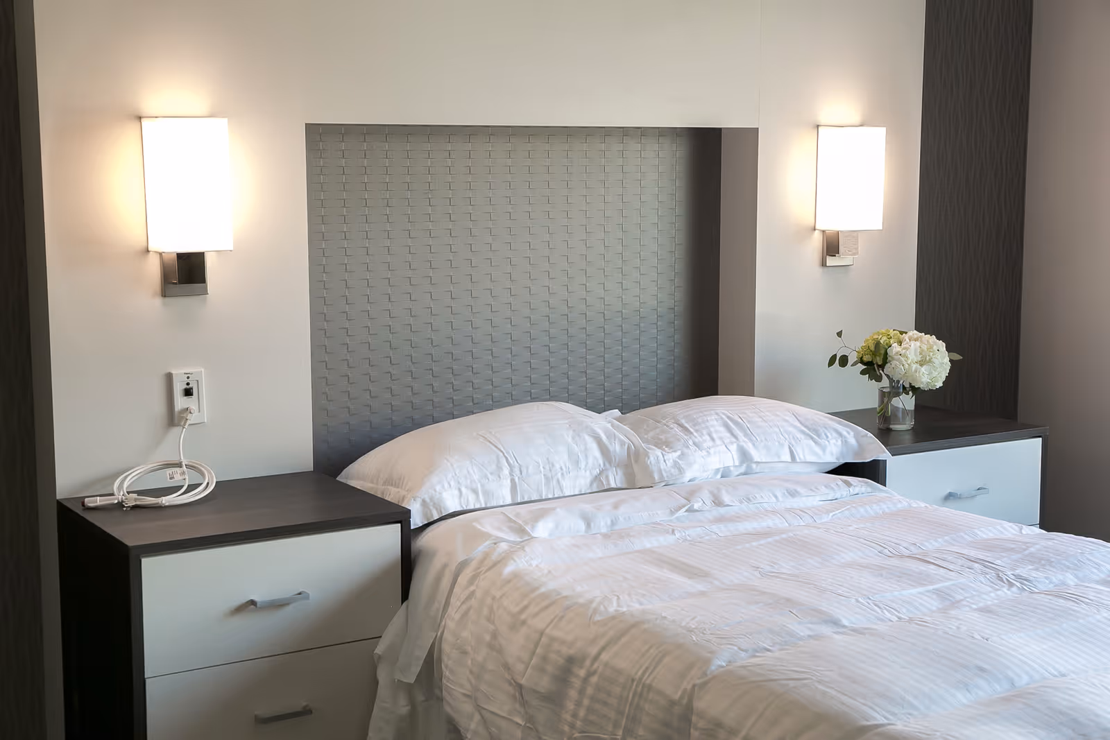A neatly made bed with white bedding and two pillows, flanked by two nightstands. Each nightstand has a modern wall-mounted light above it. One nightstand has a coiled white cable, and the other has a small vase with white flowers. The headboard has a textured gray pattern.