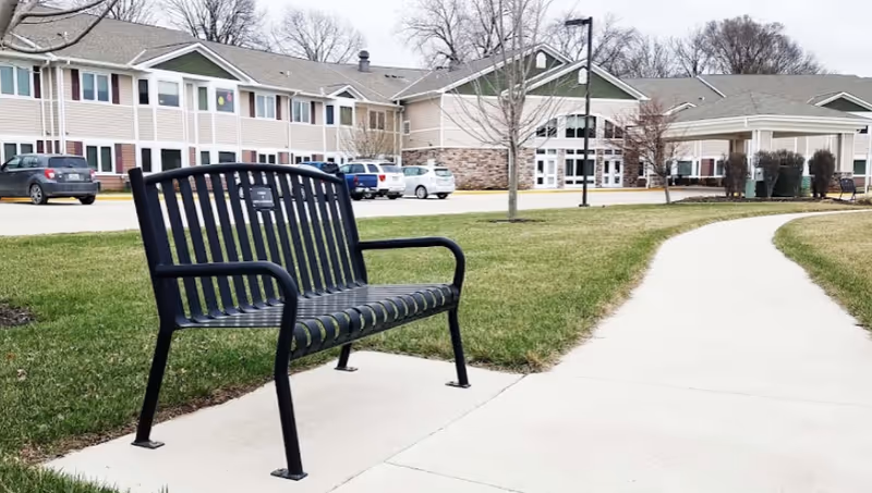 Black metal bench on a concrete sidewalk with green grass on either side, leading to a large two-story building with multiple windows and a covered entrance. Several parked cars are visible near the building, and leafless trees are scattered around the lawn.