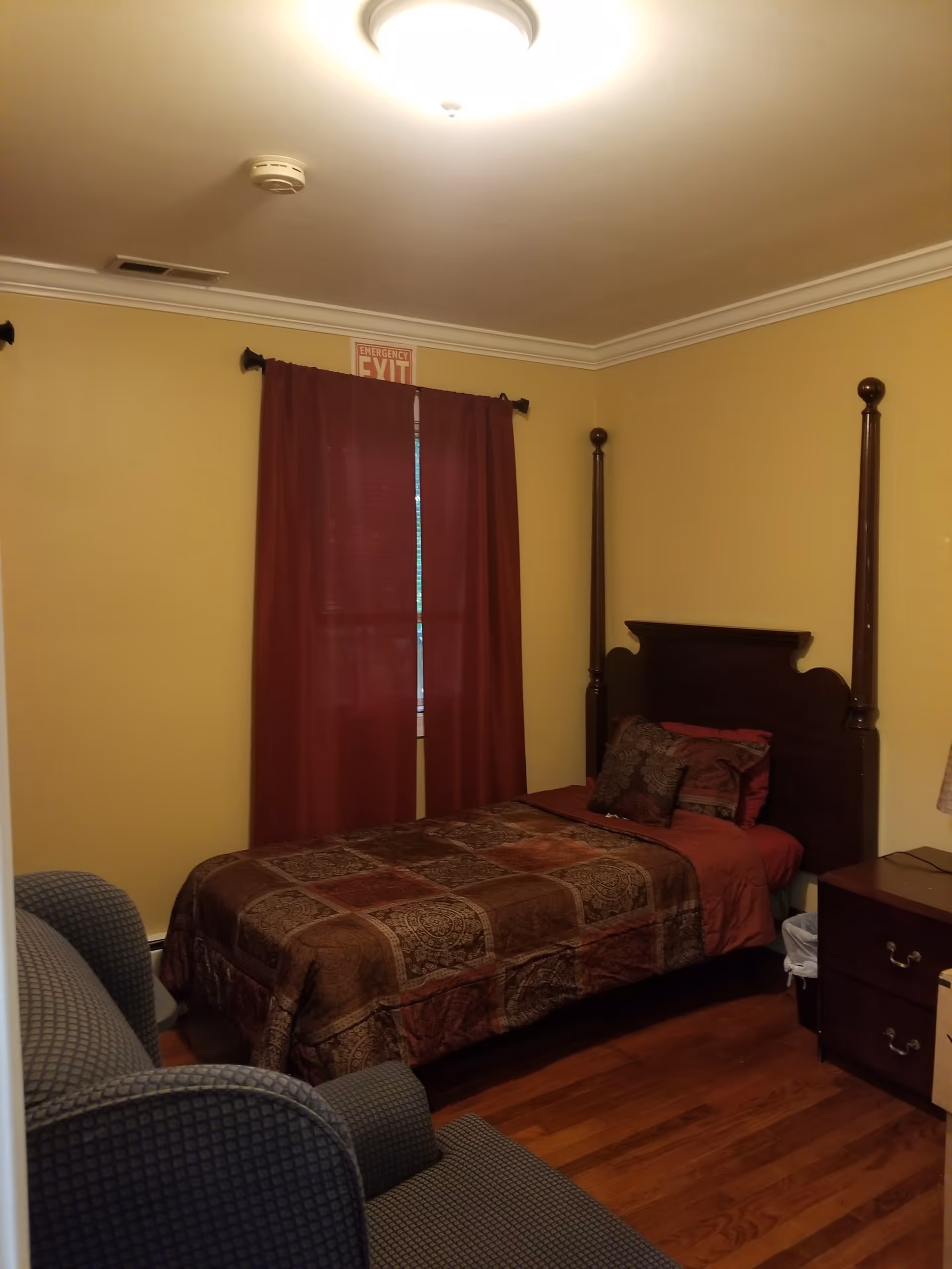 A small bedroom with yellow walls, a wooden four-poster bed with a patterned brown and red bedspread, a matching pillow, a wooden nightstand with a lamp, and a gray upholstered armchair. A window with closed red curtains is visible behind the bed, with an emergency exit sign above it.