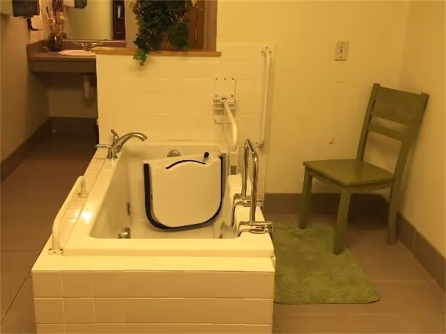 A bathroom with a walk-in bathtub featuring a side door for easy access, a faucet, and safety grab bars. There is a green chair and a green bath mat next to the tub, and a sink with a mirror is visible in the background.