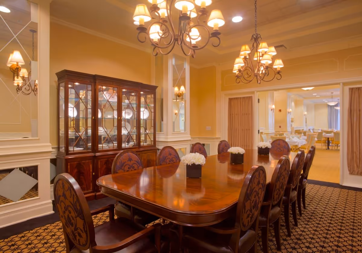 Elegant dining room with a large polished wooden table surrounded by ornate chairs. The room features a wooden china cabinet with glass doors displaying plates, multiple chandeliers with lampshades hanging from the ceiling, and wall sconces. The walls are painted in warm tones with decorative molding, and there is a patterned carpet on the floor. In the background, another dining area is visible through an open doorway.