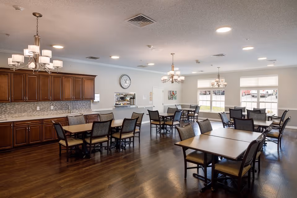 A bright dining room with multiple tables and chairs arranged neatly on a wooden floor. The room features large windows letting in natural light, wooden cabinets along one wall, and three chandeliers hanging from the ceiling. A clock and a pass-through window to a kitchen area are visible on the far wall.