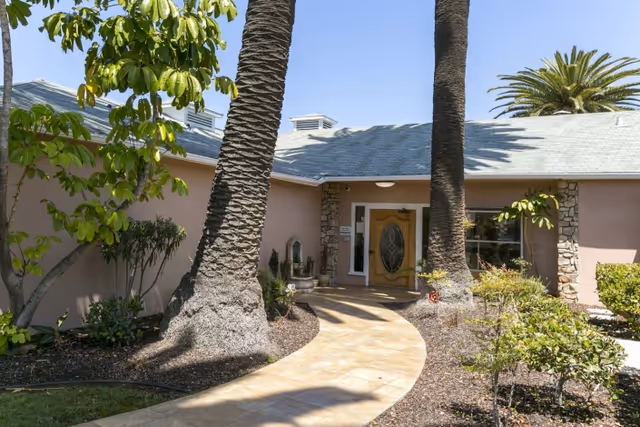Exterior view of a single-story building with a light gray roof and beige walls, featuring a wooden front door with an oval glass panel. A curved paved walkway leads to the entrance, flanked by two tall palm trees and various green shrubs and plants under a clear blue sky.