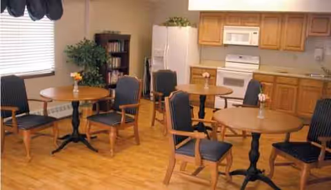 Interior dining area with round tables, chairs, and kitchenette in a senior living facility.