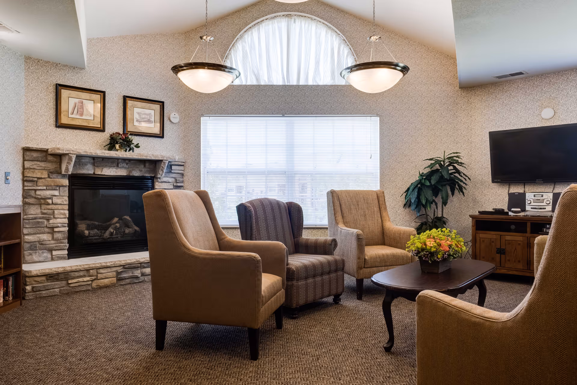 Cozy assisted-living lounge with armchairs arranged around a coffee table in front of a stone fireplace and a TV.