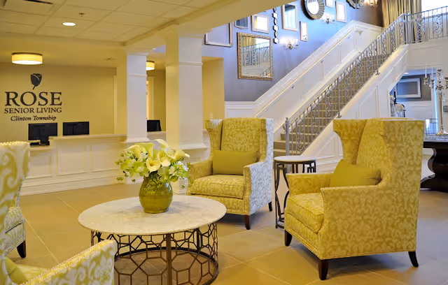 A bright and welcoming senior living facility lobby with a white reception desk, a sign reading 'ROSE SENIOR LIVING Clinton Township' on the wall, three patterned yellow armchairs around a round table with a green vase of white flowers, and a staircase with decorative railings and framed mirrors on the wall.