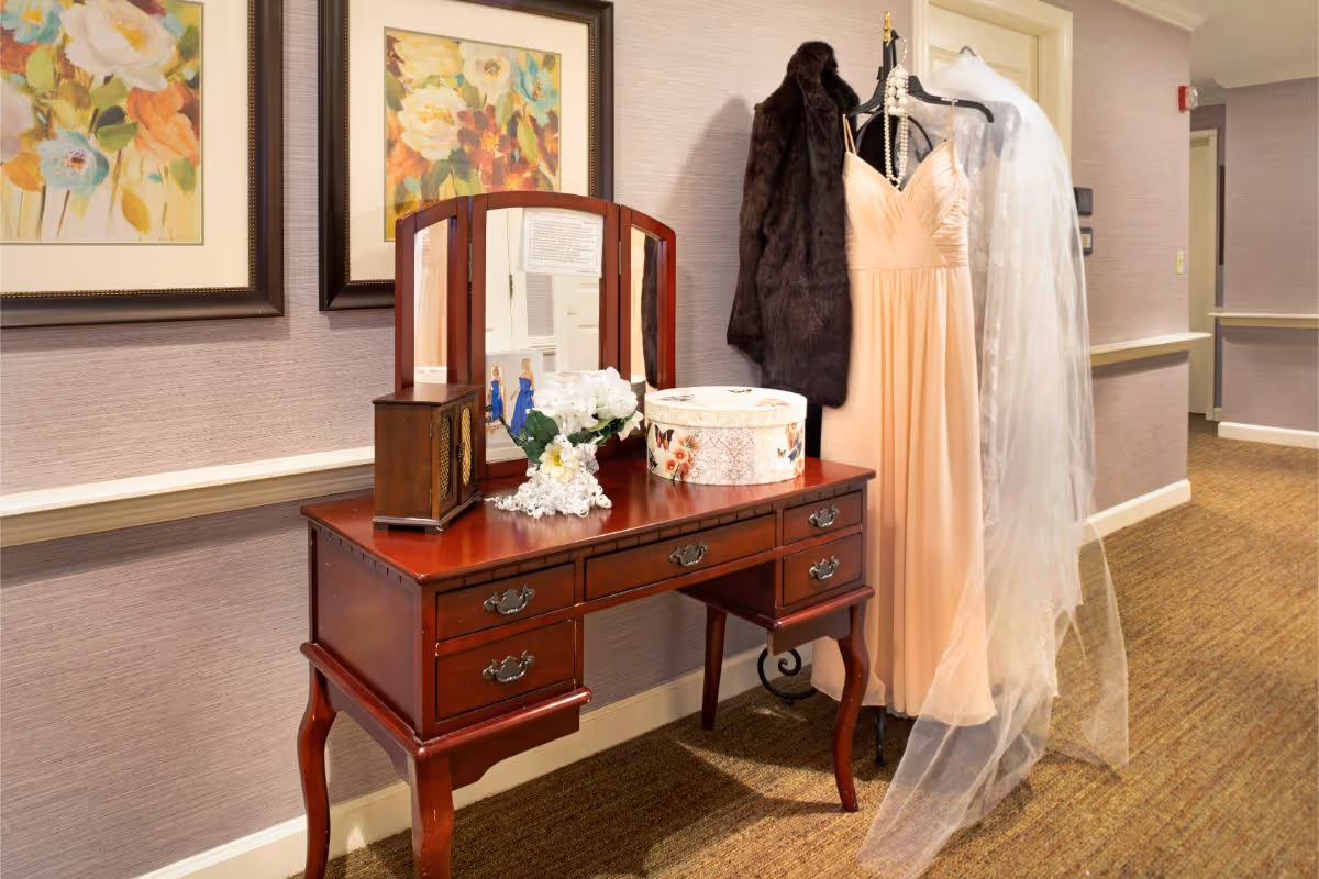 A wooden vanity table with a tri-fold mirror is placed against a wall in a hallway. On the table are a vintage radio, a bouquet of white flowers, a decorative round box, and a string of white beads. Next to the table, a dress form displays a peach-colored gown with a pearl necklace, a dark fur coat, and a long white veil. Two framed floral paintings hang on the wall above the vanity.