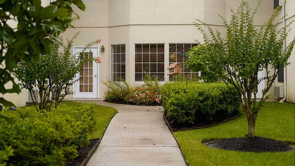 A concrete pathway leading through a well-maintained garden area with green bushes, small trees, and flowering plants, situated in front of a beige building with windows and a white door.