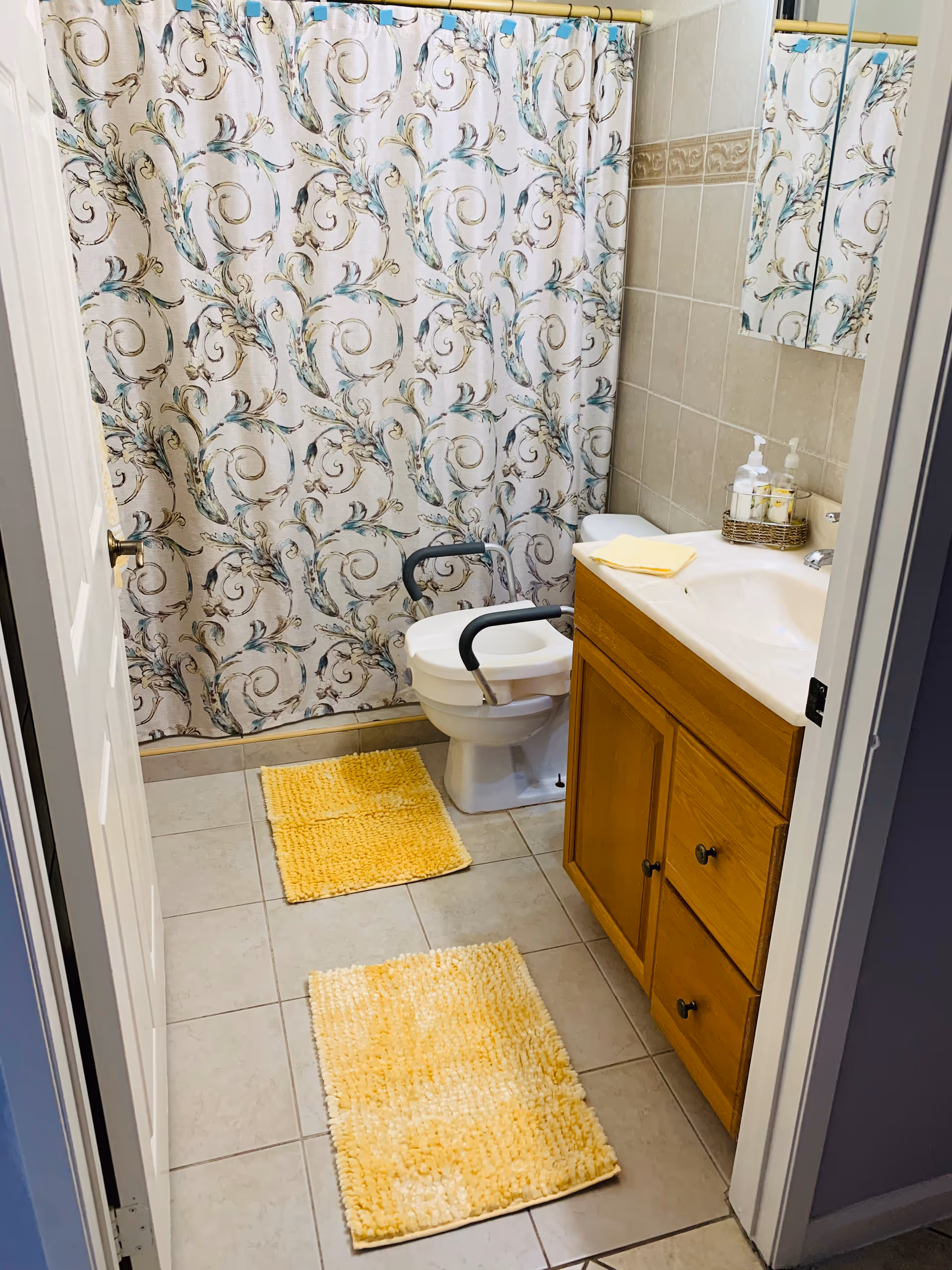 A small bathroom with beige tiled floor and walls. There is a wooden vanity with a white sink on the right side, a toilet with black safety rails in the center, and a shower with a decorative floral-patterned curtain in the background. Two yellow bath mats are placed on the floor, and a yellow towel is folded on the vanity. A mirrored medicine cabinet is mounted above the sink.
