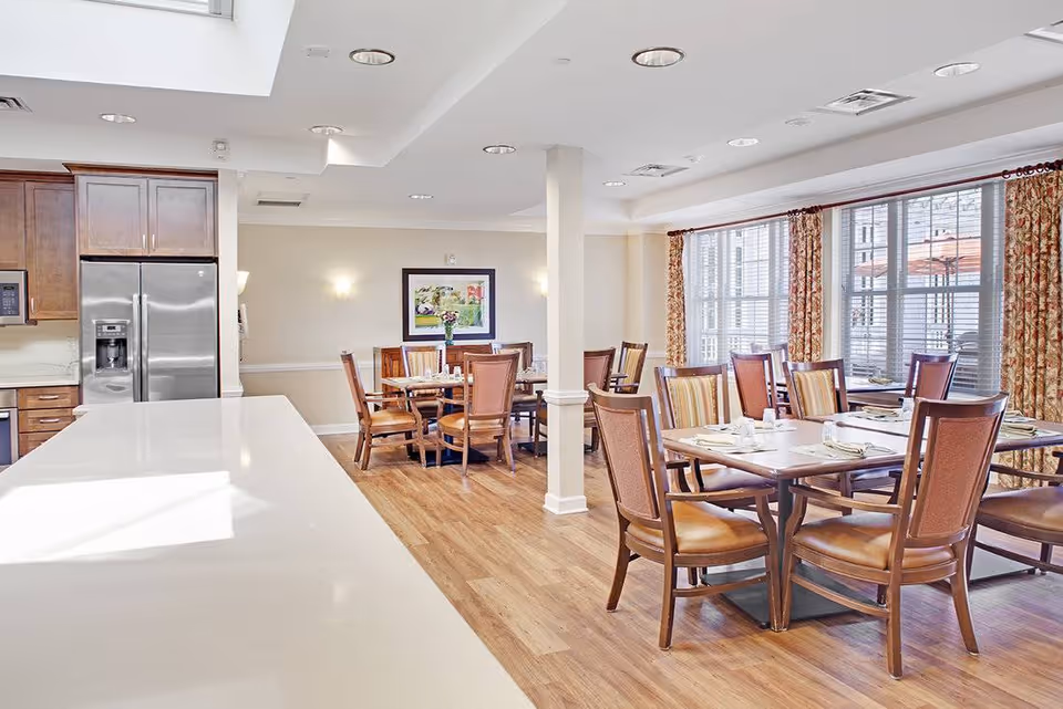 Bright dining area in a senior living facility with wooden chairs and tables set with plates and napkins. Large windows with floral curtains allow natural light to fill the room. A kitchen area with stainless steel refrigerator and wooden cabinets is visible on the left side.