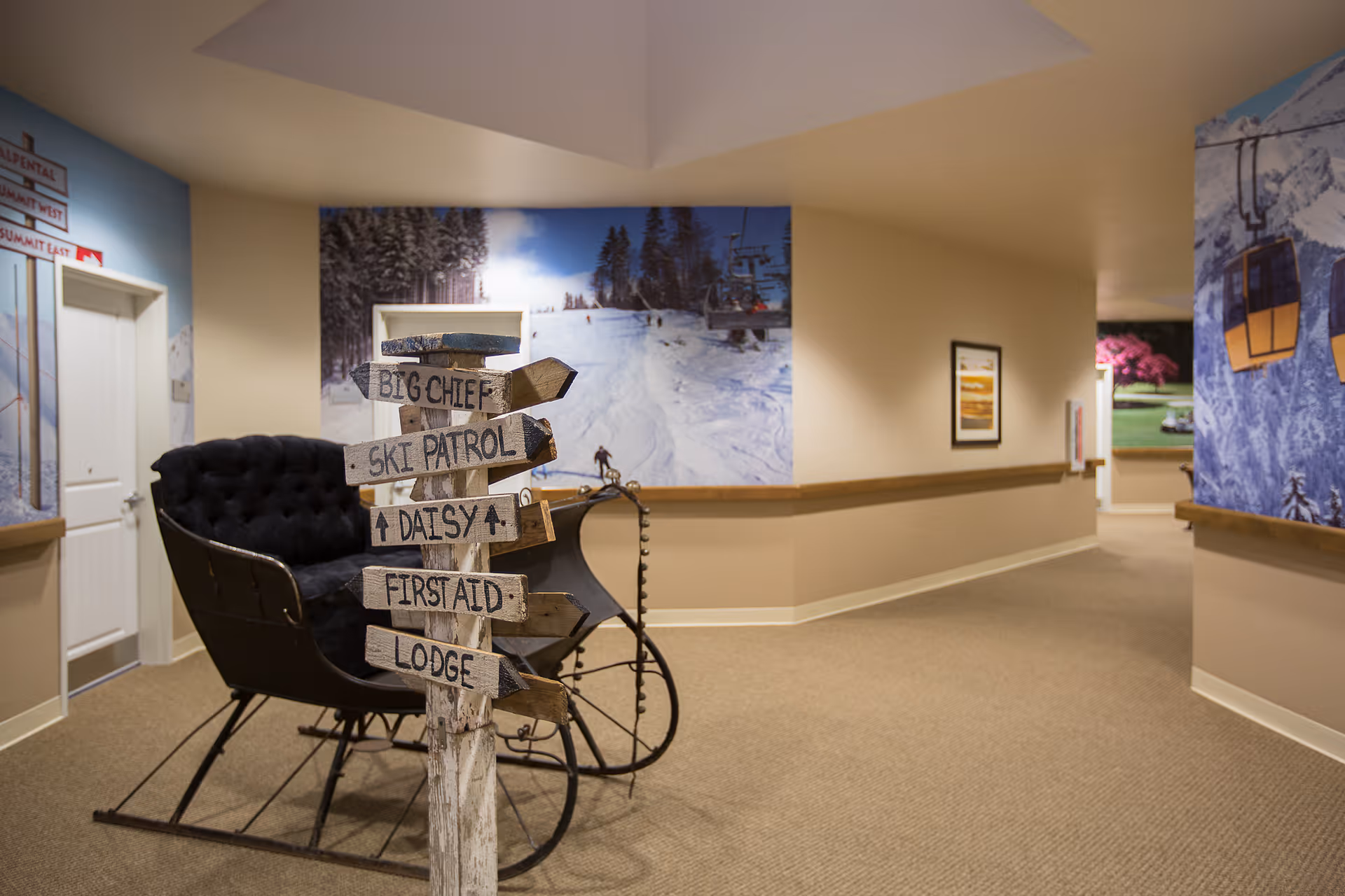 Indoor hallway with beige walls and carpeted floor featuring a vintage black sleigh and a wooden signpost with directional signs labeled Big Chief, Ski Patrol, Daisy, First Aid, and Lodge. The walls are decorated with large winter and ski-themed murals and framed pictures.
