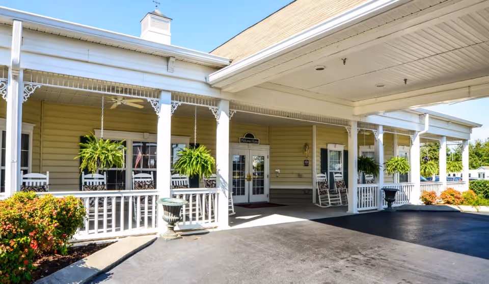 Front entrance of a senior living facility with a covered porch featuring white rocking chairs and hanging green plants. The building has beige siding and white trim, with a paved driveway in front and some landscaping with bushes and flowers.