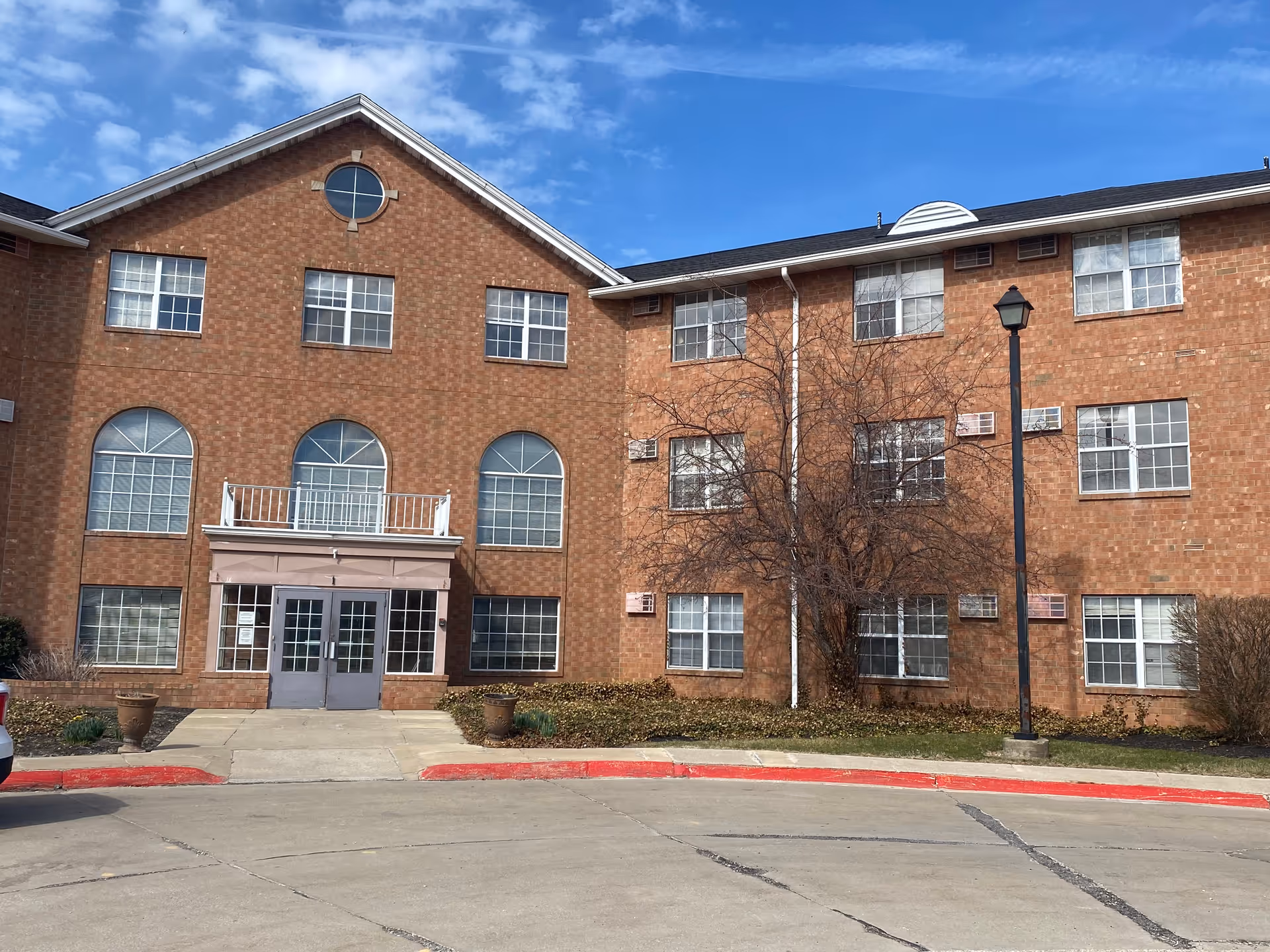 Exterior view of a three-story brick building with multiple windows and a central entrance with double glass doors. There is a small balcony above the entrance and a streetlamp in front of the building. The sky is clear and blue.