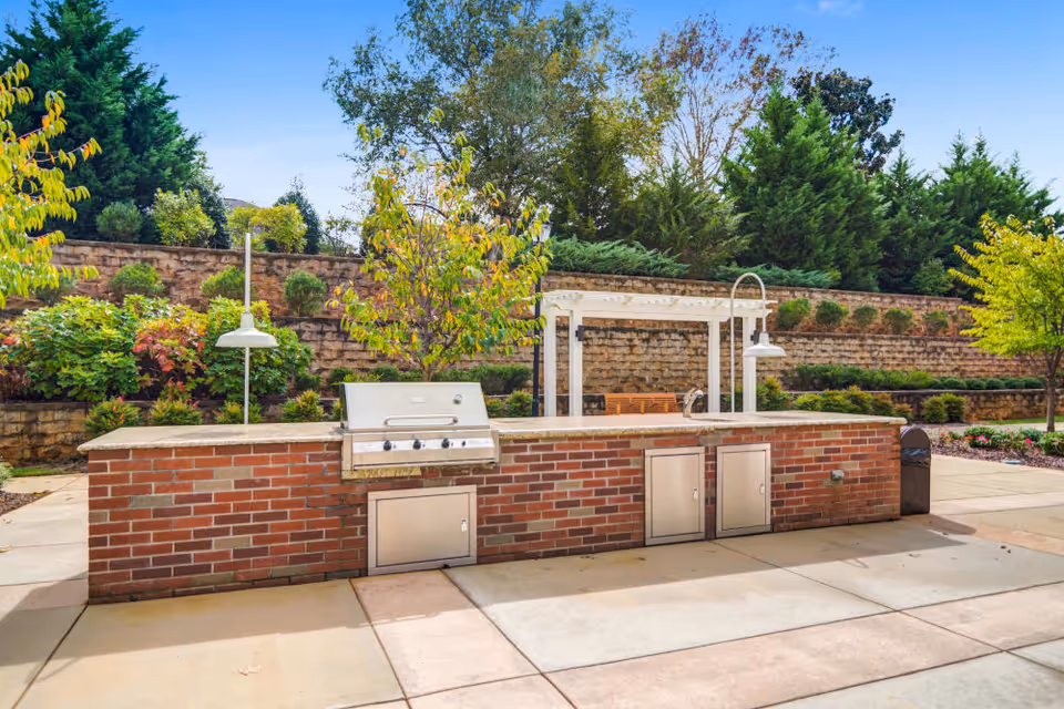 Outdoor patio area with a built-in brick barbecue grill and countertop, surrounded by landscaped greenery, trees, and a stone retaining wall under a clear blue sky.