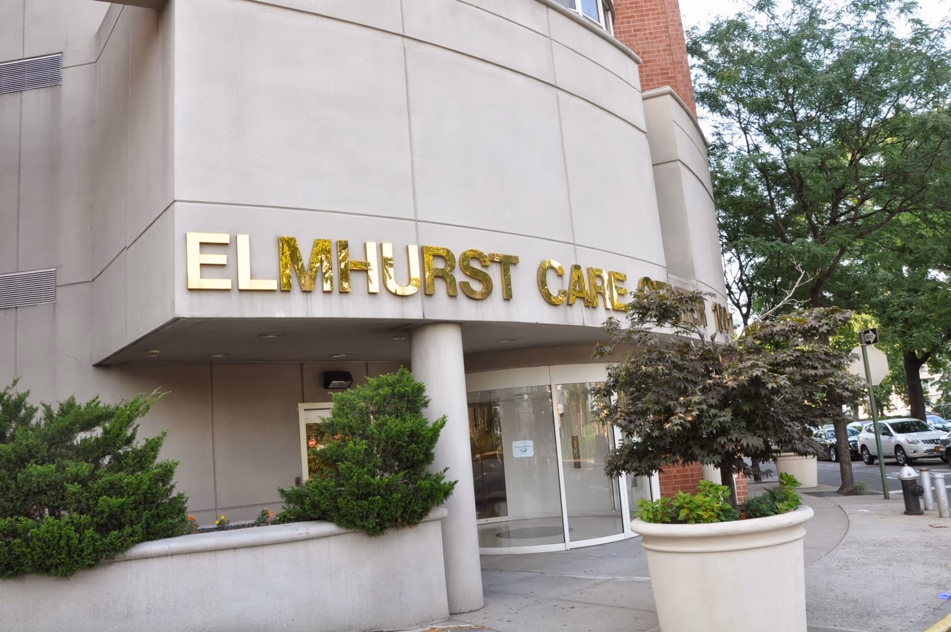 Exterior view of Elmhurst Care Center building entrance with gold letters on the facade, surrounded by greenery including bushes and a tree in a large planter, with a sidewalk and parked cars visible.