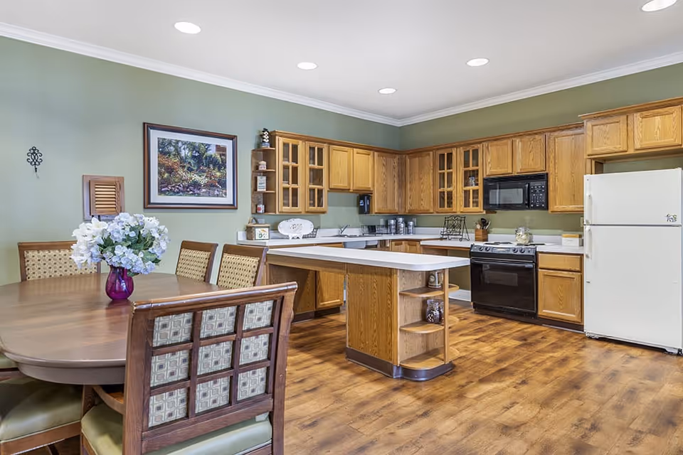 A spacious kitchen with wooden cabinets and green walls. The kitchen features a white refrigerator, black microwave, black stove, and a central island with open shelving. To the left, there is a wooden dining table with six cushioned chairs and a vase of white flowers. A framed painting and decorative items are displayed on the walls and shelves.