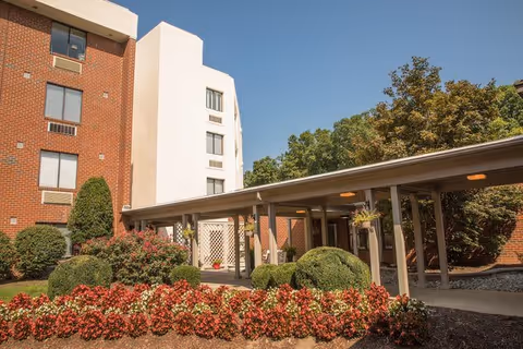 Entrance of a brick and white senior living building with a covered walkway and landscaped flower beds.