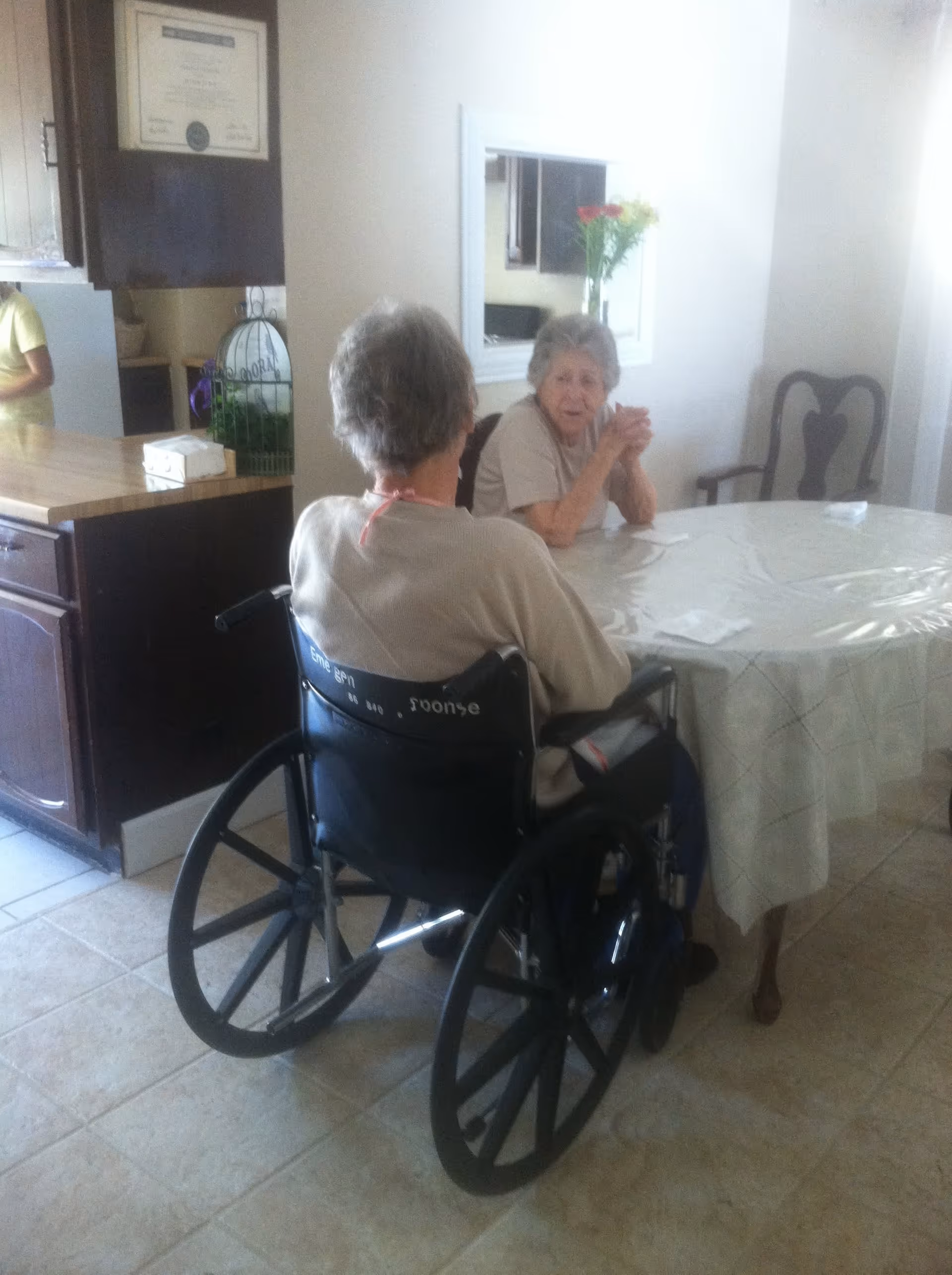 Two elderly women sitting at a dining table in a room with tiled floor. One woman is in a wheelchair facing away from the camera, and the other woman is sitting across from her, looking towards the camera. The room has a kitchen counter with cabinets, a framed certificate on the wall, a mirror, and a vase with flowers on the table.