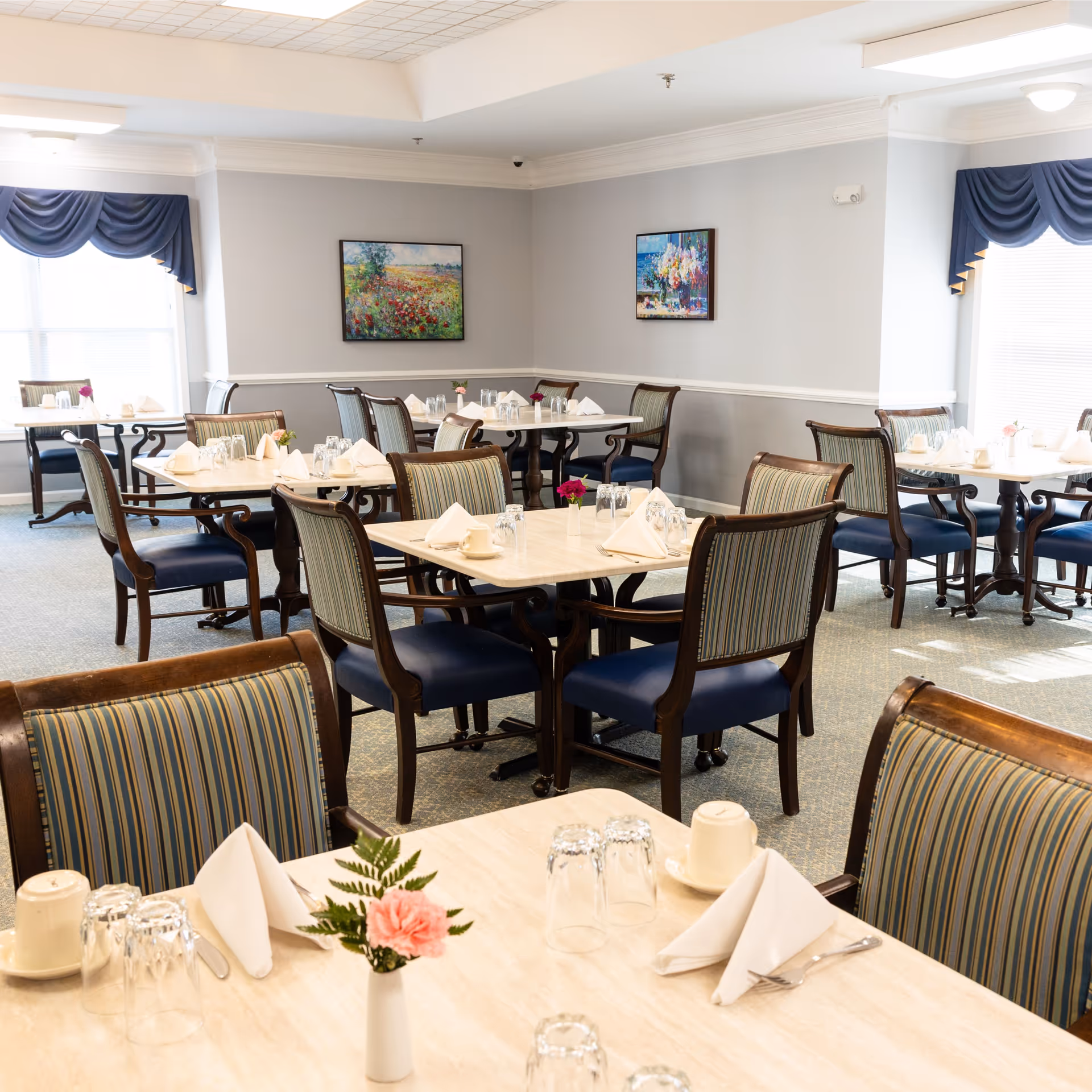 A bright dining room with multiple tables set with white napkins, upside-down glasses, cups, and small flower vases. The room has striped upholstered chairs, light-colored walls with two framed paintings, and windows with blue valances allowing natural light to enter.