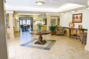 A spacious and well-lit senior living facility common area with tiled floors, a round wooden table with a vase of white flowers in the center, two upholstered chairs, a wooden console table with decorative items, and framed artwork on the walls. In the background, there is a dining area with tables and chairs visible through an open doorway.