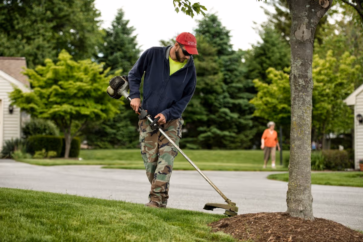 A landscaper wearing a red cap trims grass with a string trimmer near a tree in a residential community while a person walks in the background.