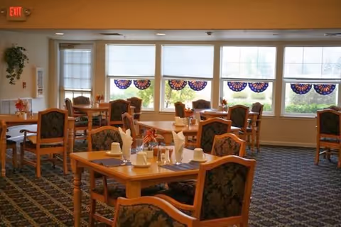 Dining room with wooden tables and upholstered chairs set with place settings beside large windows.