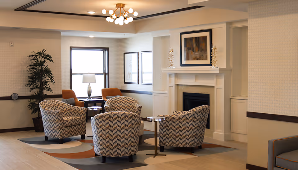 A cozy senior living common area with four patterned armchairs arranged around a small round table on a multicolored rug. There is a fireplace with a framed abstract painting above it, two decorative candle holders on the mantel, a potted plant in the corner, and a window with a table lamp and two orange chairs nearby. The room has soft lighting from a modern ceiling fixture.