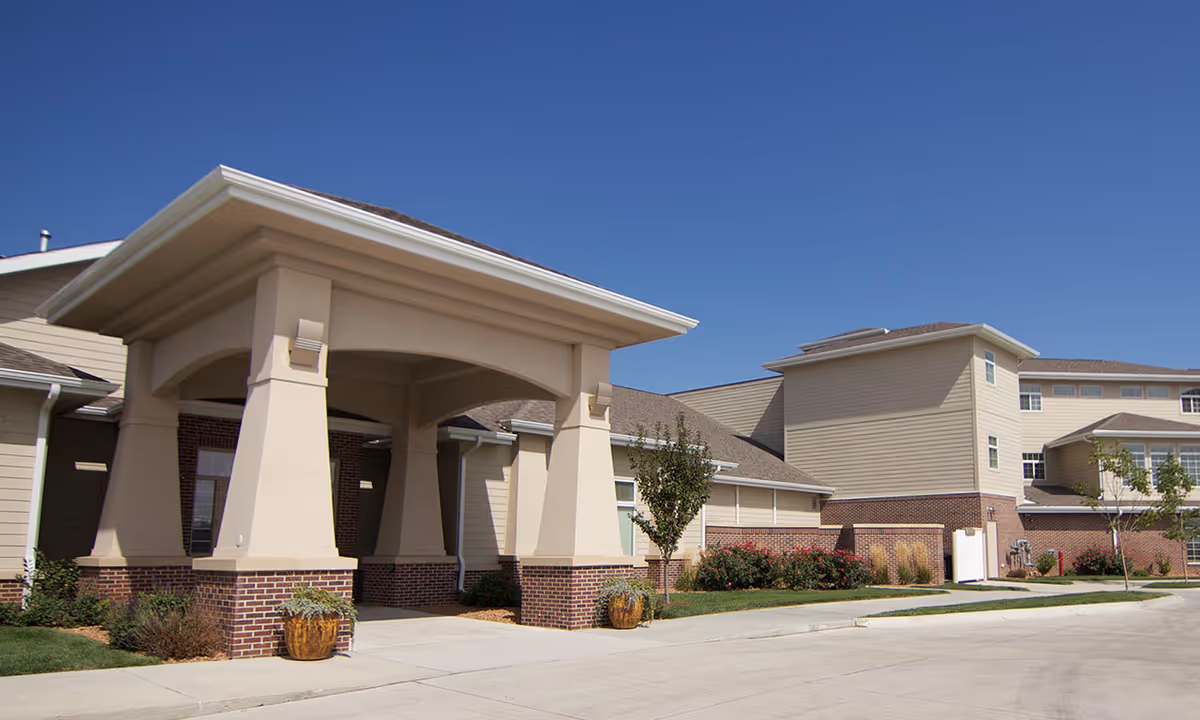 Exterior view of Whispering Creek Senior Living facility showing a covered entrance with large beige columns on brick bases, surrounded by landscaped greenery and a clear blue sky.