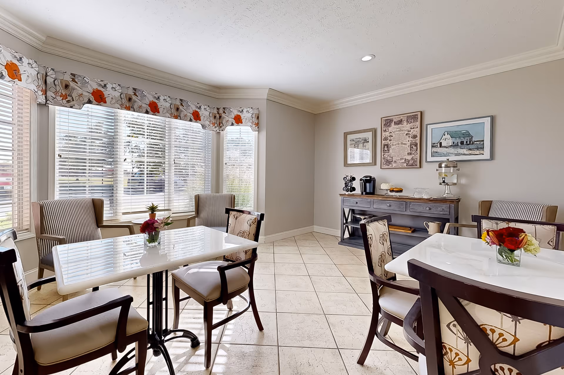 Bright assisted‑living dining room with two white tables, upholstered chairs, a large bay window with blinds and floral valance, and a sideboard with framed artwork.