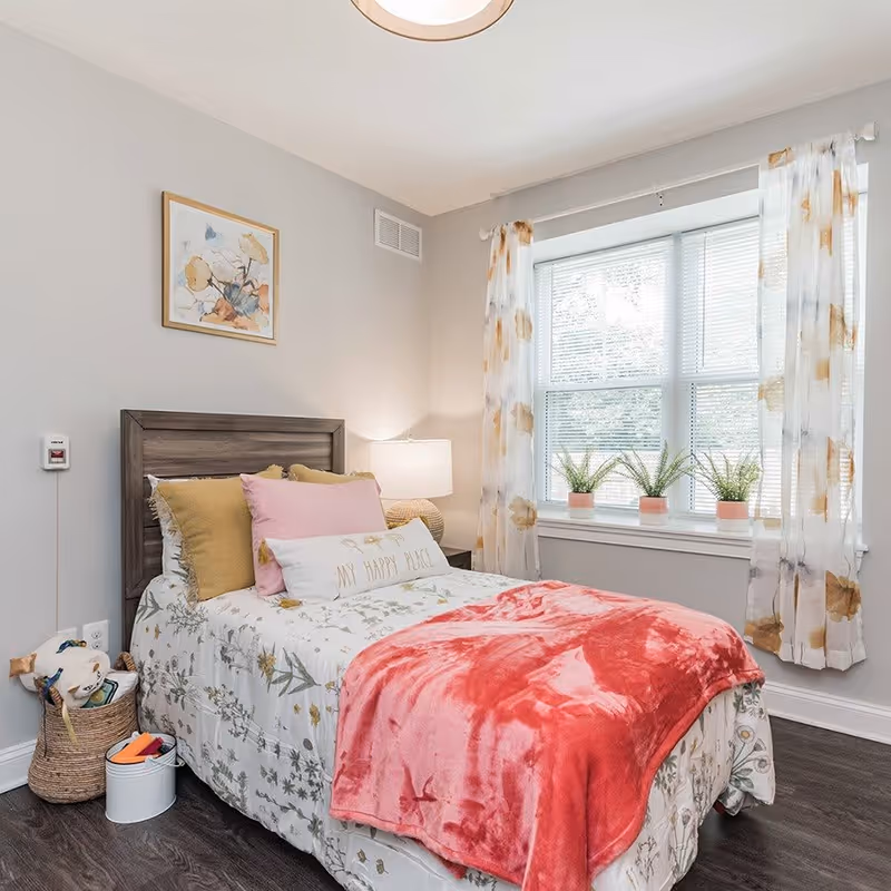 A cozy bedroom with a single bed featuring floral bedding, a pink throw blanket, and multiple pillows including one that says 'MY HAPPY PLACE'. There is a wooden headboard, a bedside table with a lamp, a window with floral curtains, and three small potted plants on the windowsill. A basket and a small white container are on the floor next to the bed.