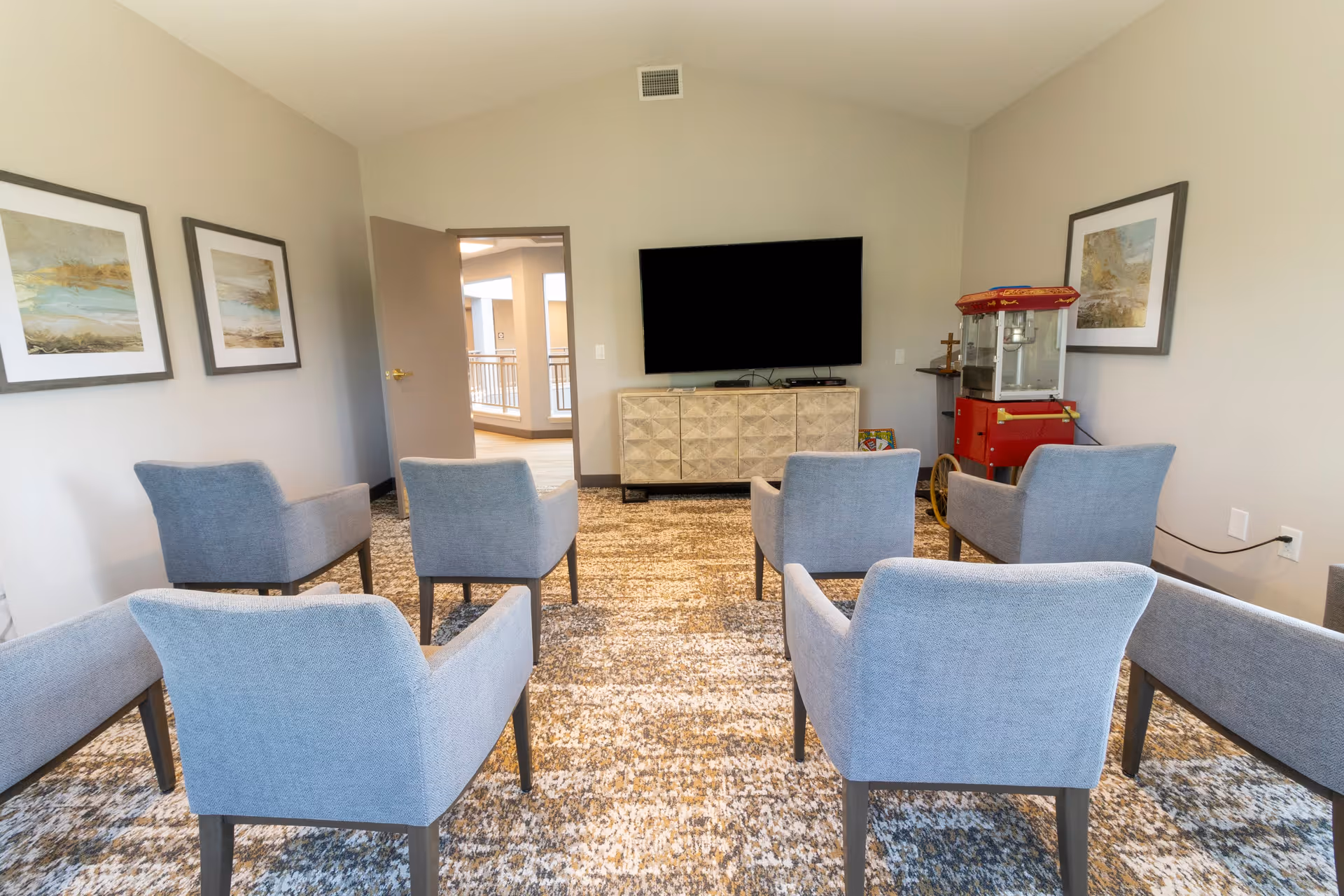 A small lounge area with six gray upholstered chairs arranged in two rows facing a wall-mounted flat screen TV. Below the TV is a light-colored cabinet. To the right of the TV is a red popcorn machine. The room has beige walls with three framed landscape paintings and a patterned carpet. An open door leads to a hallway with railings and additional rooms.