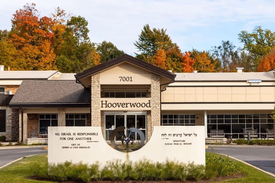 Front entrance of the Hooverwood Living building showing the main entry with address 7001, a stone commemorative sign, and autumn trees in the background.